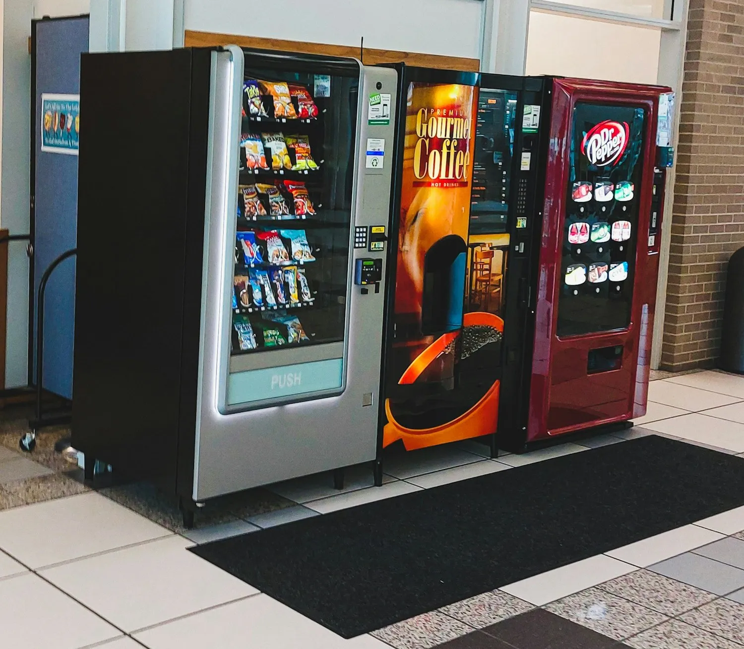 A snack vending machine beside a coffee machine and a drinks machine in a building lobby. Illustrative photo, not MVend machines.